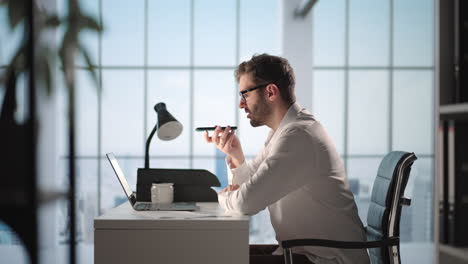 Young-Guy-Talking-On-Mobile-Phone-And-Taking-Notes-Using-Voice-Assistant-Writing-In-Notebook.-Smiling-Man-Sitting-At-Desk-With-Laptop-At-Home-Office